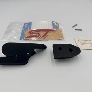 May include: A black automotive grille emblem mount set including a red and silver ST badge, two small metal screws, and an alcohol prep pad. The components are arranged on a plain surface, featuring a textured black plastic bracket and a separate mounting piece.