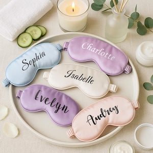 May include: A collection of personalized satin sleep masks in pastel colors, each embroidered with a name: Sophia, Charlotte, Isabelle, Evelyn, and Audrey. The image includes a lit candle, a small white towel, and skincare products.