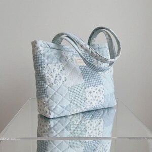 May include: A quilted tote bag in shades of light blue and white. The bag features a patchwork design with floral and gingham patterns. The handles are also quilted. The bag is sitting on a clear surface.