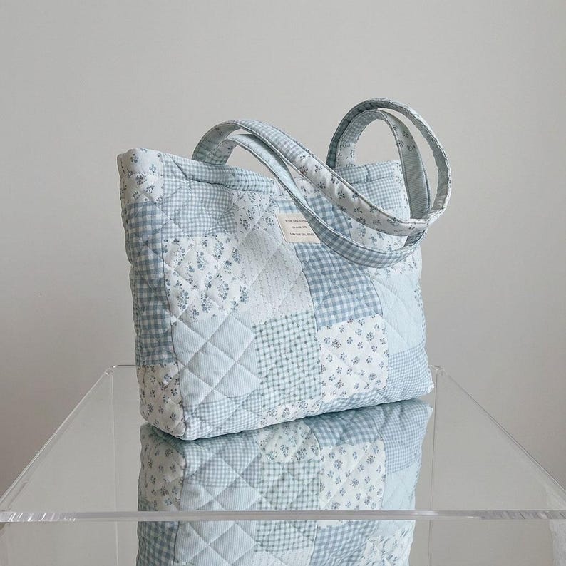 May include: A quilted tote bag in shades of light blue and white. The bag features a patchwork design with floral and gingham patterns. The handles are also quilted. The bag is sitting on a clear surface.