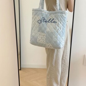 May include: A light blue quilted tote bag with a patchwork design. The bag features a variety of patterns, including floral and gingham, and the name "Stella" embroidered in a cursive font. The bag has matching handles.