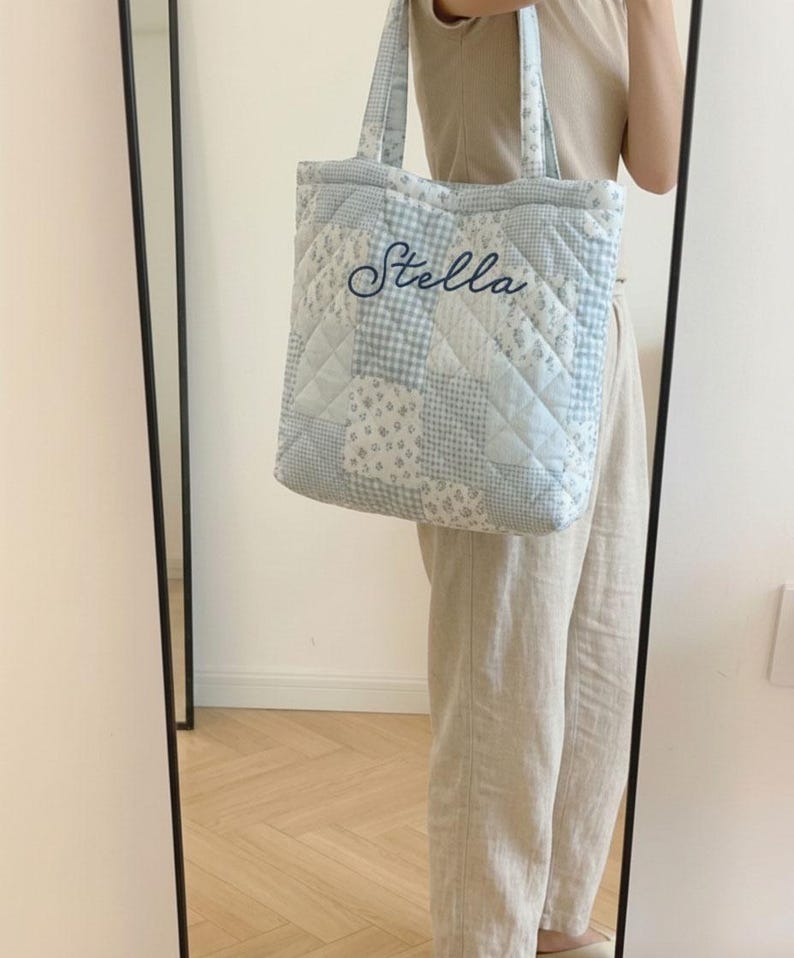 May include: A light blue quilted tote bag with a patchwork design. The bag features a variety of patterns, including floral and gingham, and the name "Stella" embroidered in a cursive font. The bag has matching handles.