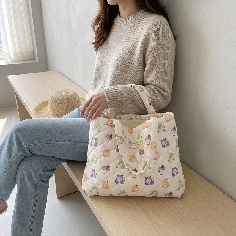 May include: Cream-colored quilted tote bag with a repeating pattern of cartoon characters in various colors. The bag has two handles and a small label. The bag is held by a person wearing a beige sweater and blue jeans.