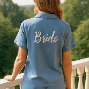 Op de afbeelding: Lichtblauwe pyjamaset met witte bies, bestaande uit een top met korte mouwen en een short. Het woord "Bride" is in het wit op de achterkant van de top gedrukt. De persoon staat op een balkon met uitzicht op een groen landschap.