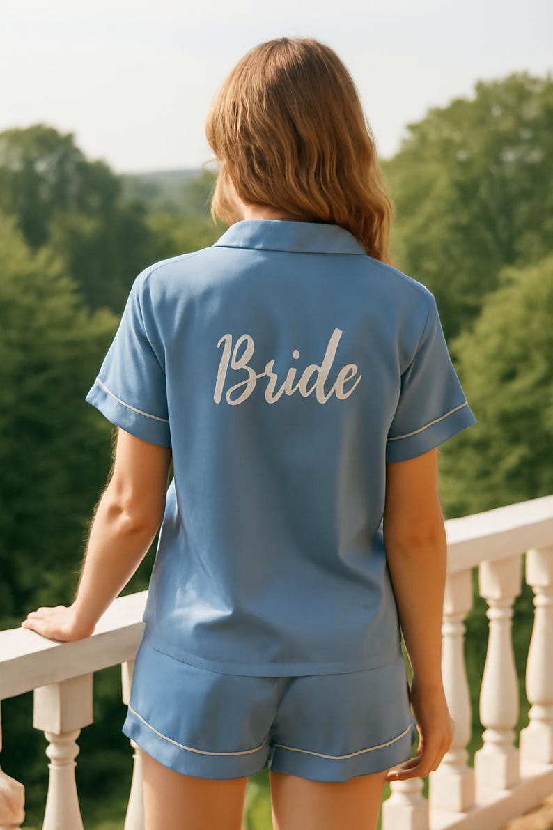 Op de afbeelding: Lichtblauwe pyjamaset met witte bies, bestaande uit een top met korte mouwen en een short. Het woord "Bride" is in het wit op de achterkant van de top gedrukt. De persoon staat op een balkon met uitzicht op een groen landschap.
