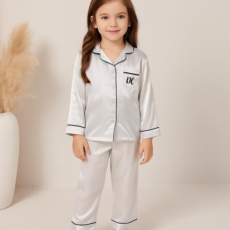 May include: A white satin pajama set with black trim and the embroidered initials "DC" on the pocket. The long-sleeved top has a button-down front and the pants are full-length. The pajamas are designed for children.