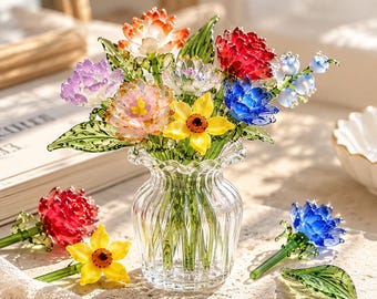 May include: A clear, fluted glass vase holds a bouquet of handcrafted glass flowers in vibrant colors including red, blue, yellow, purple, and white. Additional loose glass flowers and a green glass leaf are placed on the surface nearby.