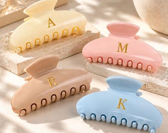 May include: Four plastic hair claw clips in pastel colours: cream, pink, beige, and light blue. Each clip features a gold-coloured letter embossed on the centre: A on the cream clip, M on the pink clip, R on the beige clip, and K on the blue clip.