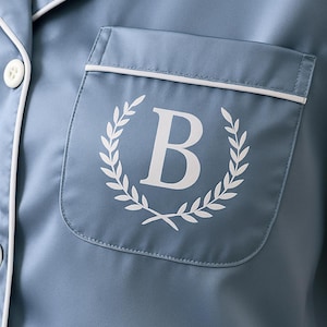 Op de afbeelding: Close-up van een blauwe pyjamatop met witte bies en een zak. De zak heeft een wit laurierkransontwerp rond een grote witte letter "B". De pyjamatop heeft witte knopen.