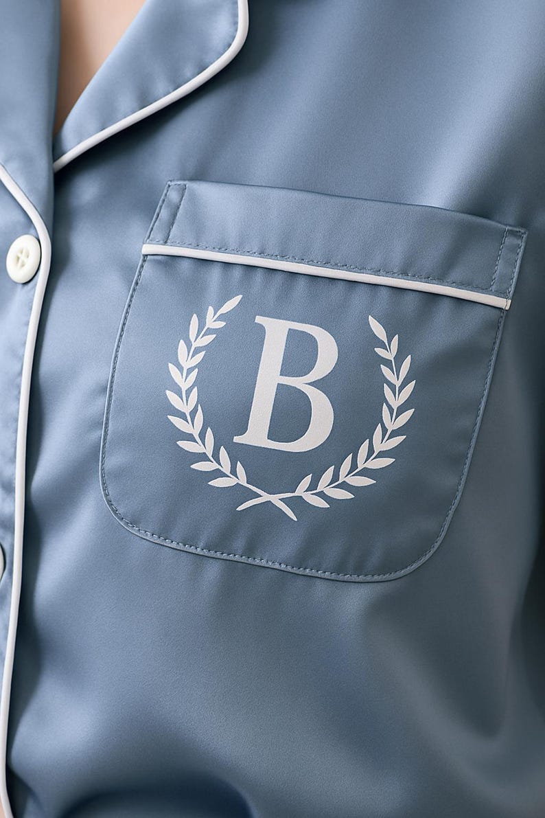Op de afbeelding: Close-up van een blauwe pyjamatop met witte bies en een zak. De zak heeft een wit laurierkransontwerp rond een grote witte letter "B". De pyjamatop heeft witte knopen.