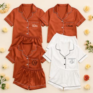May include: Matching pajama sets in rust and white. The rust-colored sets have white trim and personalized text that reads "Kita" and "Olivia". The white set has black trim and the initials "ABC". Each set includes a short-sleeved top and shorts.