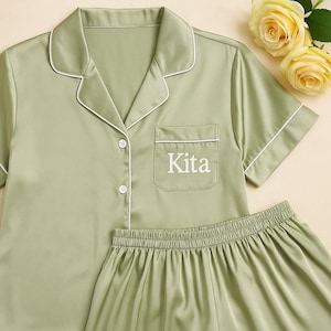 Op de afbeelding: Saliegroene pyjamaset met witte bies. De top met korte mouwen heeft een borstzak met de naam "Kita" in het wit geborduurd. De set bevat een bijpassende short. Twee gele rozen staan op de achtergrond.