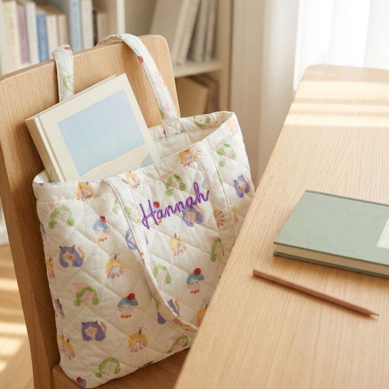 May include: A white quilted tote bag with a colorful pattern of cartoon characters and the name "Hannah" embroidered in purple. The bag is filled with a book and sits on a wooden chair. A book and pencil are on a wooden table.