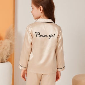 May include: A beige satin pajama set with long sleeves and pants. The back of the top features the script "Flower girl" in black. The edges of the top and sleeves are trimmed with black piping. The pajama set is designed for children.