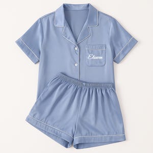 Op de afbeelding: Een lichtblauwe pyjamaset met witte biezen. De top met korte mouwen heeft een borstzak met het woord "Elawen" geborduurd. De bijpassende shorts hebben een elastische tailleband. De set is gemaakt van een gladde, zijdeachtige stof.