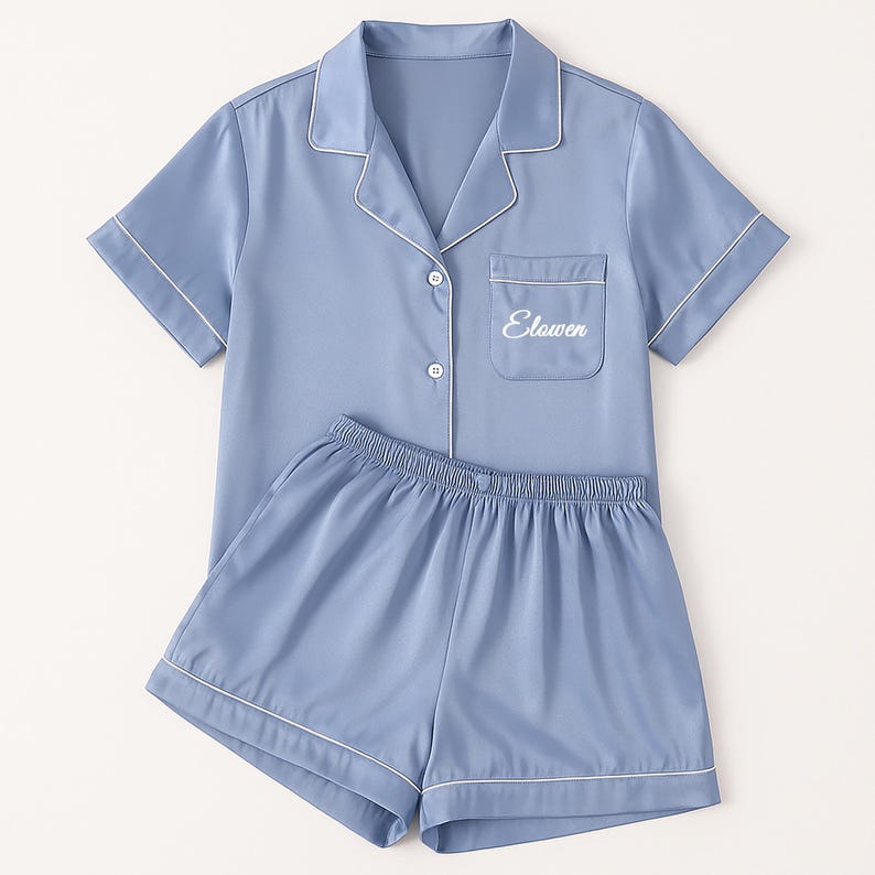 Op de afbeelding: Een lichtblauwe pyjamaset met witte biezen. De top met korte mouwen heeft een borstzak met het woord "Elawen" geborduurd. De bijpassende shorts hebben een elastische tailleband. De set is gemaakt van een gladde, zijdeachtige stof.