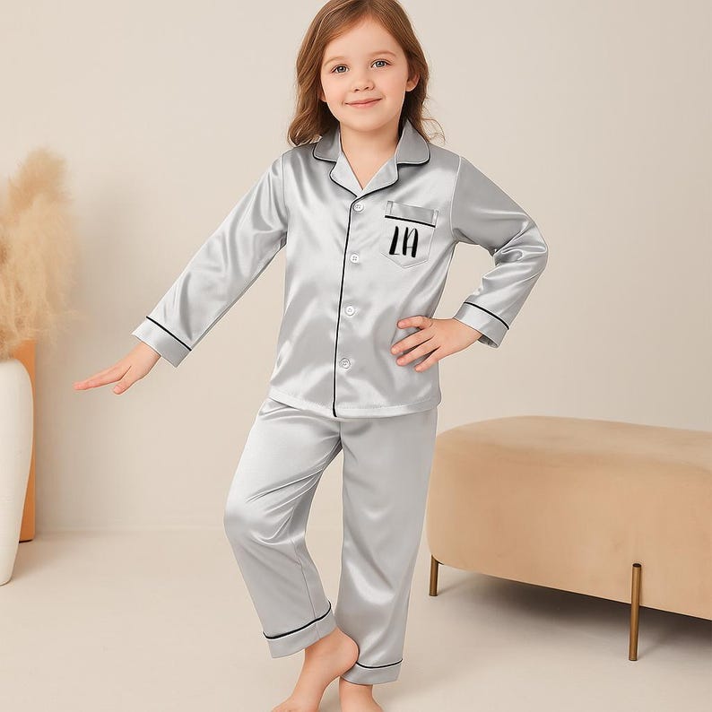 May include: A child wearing a silver satin pajama set. The long-sleeved top has black trim and the letter "In" on the pocket. The matching pants have a relaxed fit. The child is smiling and standing in front of a neutral background.