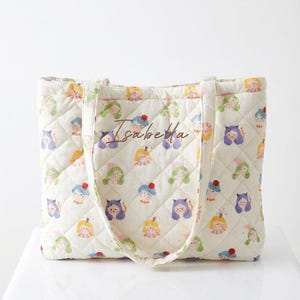 May include: Cream-colored quilted tote bag featuring a repeating pattern of whimsical cartoon characters in various colors. The bag has a diamond-stitched pattern and the name "Isabella" is written on the front.