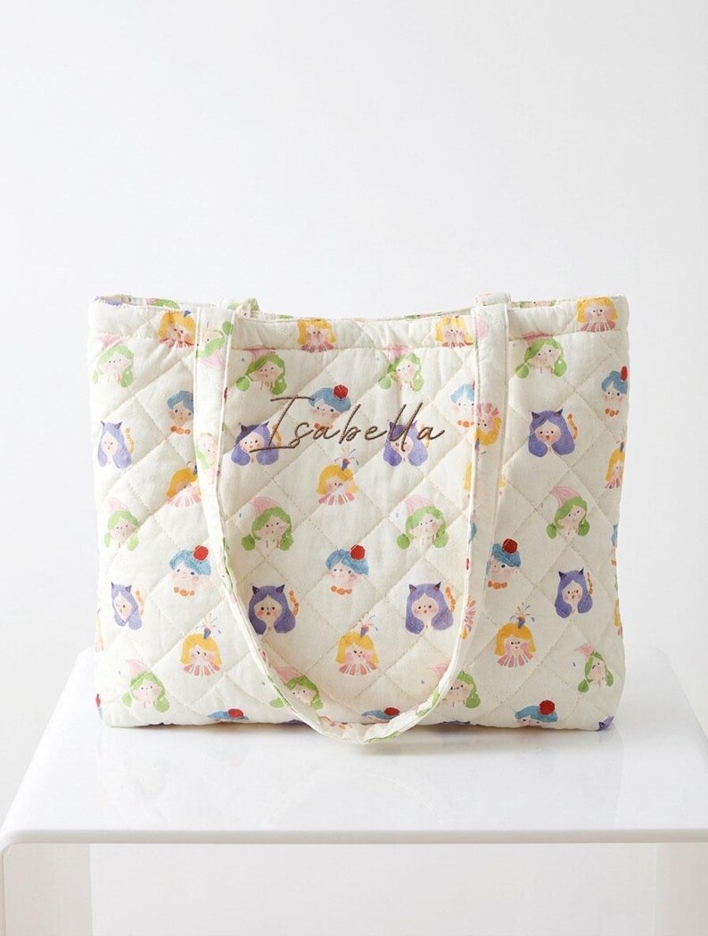 May include: Cream-colored quilted tote bag featuring a repeating pattern of whimsical cartoon characters in various colors. The bag has a diamond-stitched pattern and the name "Isabella" is written on the front.