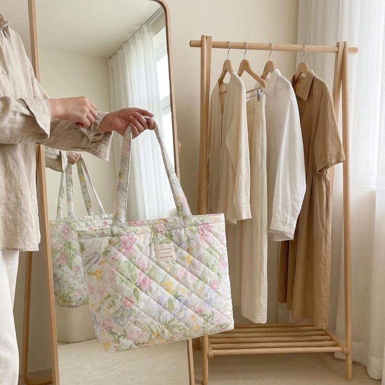 May include: A quilted tote bag with a floral pattern in pastel colors, held in front of a mirror. The bag has a diamond pattern and a small label. Clothing hangs on a wooden rack in the background.