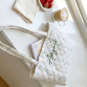 May include: A quilted tote bag with floral patterns and the name "Abigail" embroidered in green. The bag contains a notebook with cartoon illustrations. A plate of strawberries, a cup of coffee, and a folded cloth are also visible.
