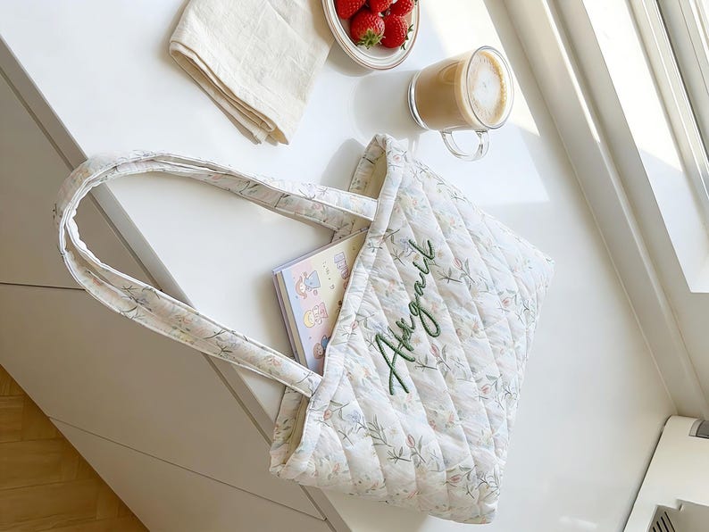May include: A quilted tote bag with floral patterns and the name "Abigail" embroidered in green. The bag contains a notebook with cartoon illustrations. A plate of strawberries, a cup of coffee, and a folded cloth are also visible.