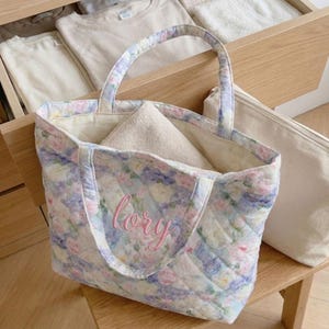 May include: Quilted tote bag with a floral pattern in pastel colors, featuring pink, blue, and white flowers. The bag has a matching handle and the name "lory" embroidered in pink. A folded beige towel is inside the bag.