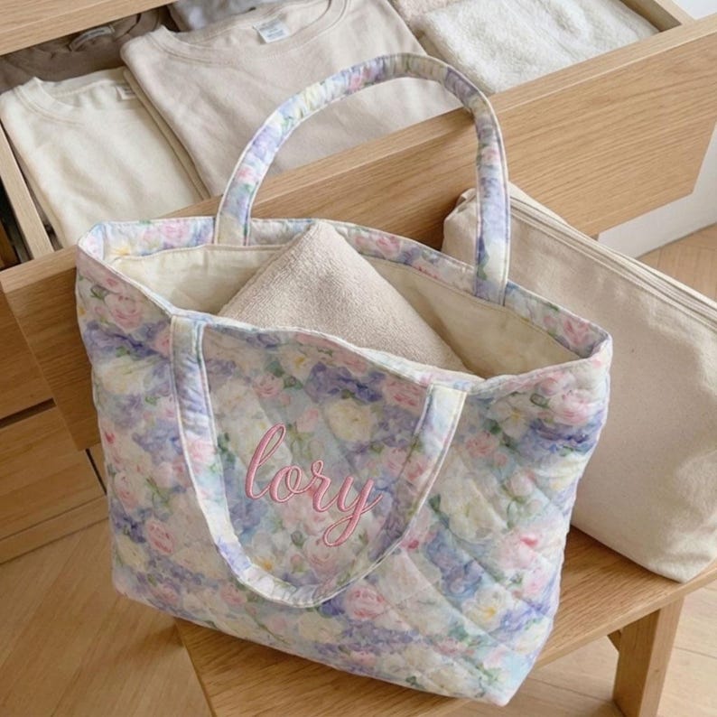 May include: Quilted tote bag with a floral pattern in pastel colors, featuring pink, blue, and white flowers. The bag has a matching handle and the name "lory" embroidered in pink. A folded beige towel is inside the bag.