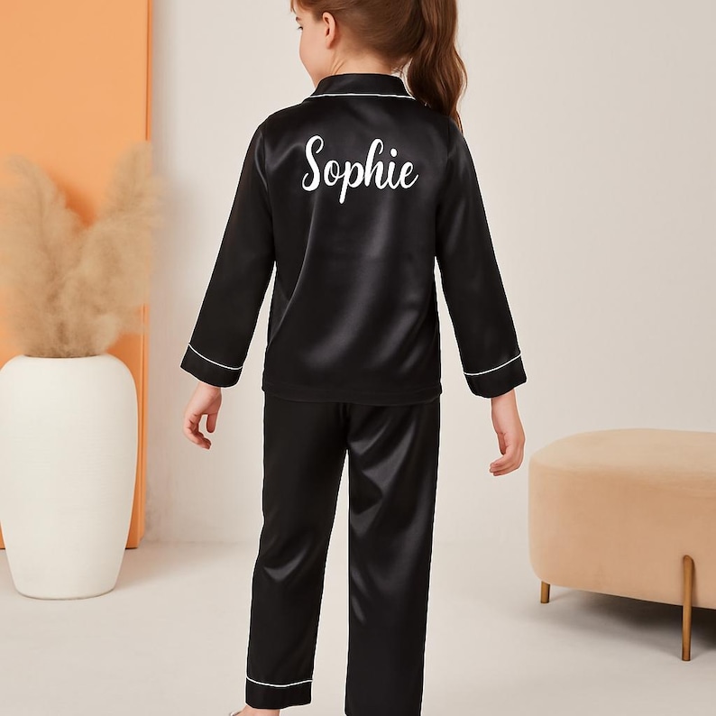 May include: Black satin pajama set with white trim. The top features long sleeves and the name "Sophie" in white script on the back. The pajama pants are long and have white trim at the ankles.