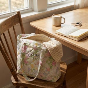 May include: A floral quilted tote bag with a zipper closure, sitting on a wooden chair. The bag features a pastel floral pattern with pink, yellow, and green hues. A beige knit hat is partially visible inside the bag. A mug, keys, and a notebook are on a wooden table.
