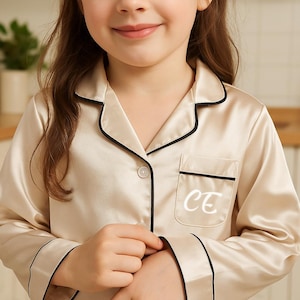 May include: Cream-colored pajama set with black trim. The top has a pocket with the white initials "CE". The long-sleeved top has a button-down front. The pajamas are made of a silky material.