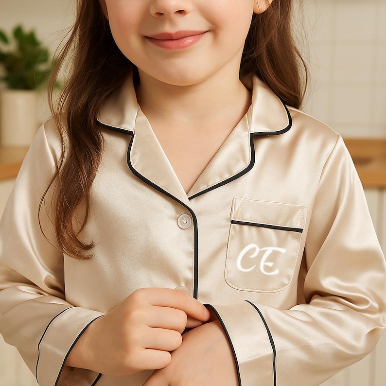 May include: Cream-colored pajama set with black trim. The top has a pocket with the white initials "CE". The long-sleeved top has a button-down front. The pajamas are made of a silky material.