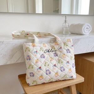 May include: Cream-colored quilted tote bag with a repeating pattern of cartoon characters in various colors. The bag has a diamond pattern and the name "Chloe" is embroidered on the front. The bag is sitting on a wooden stool.