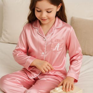 May include: Pink satin pajama set with white trim, featuring a button-down top with a pocket and the letter "A" embroidered on it. The set includes matching pants. A small gift box with a ribbon is also present.
