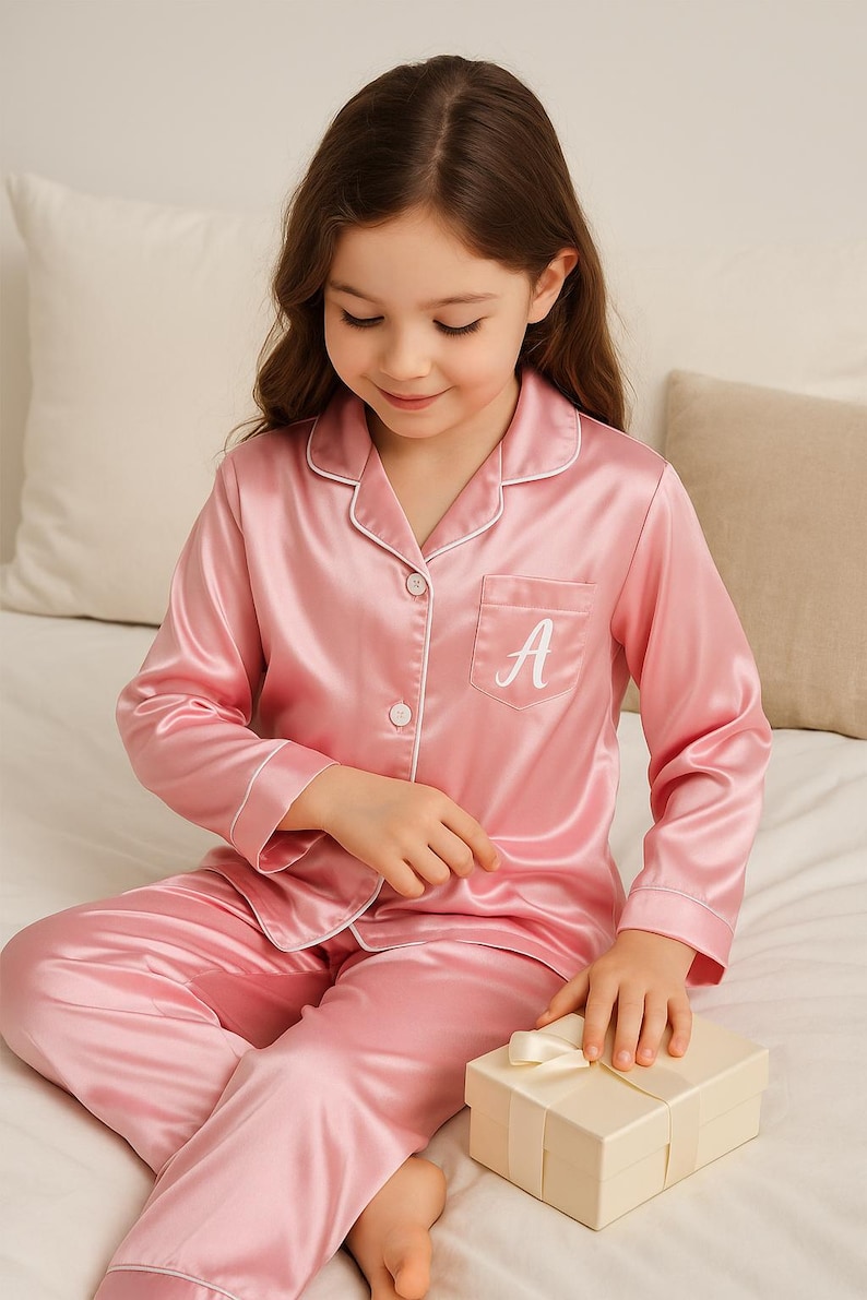 May include: Pink satin pajama set with white trim, featuring a button-down top with a pocket and the letter "A" embroidered on it. The set includes matching pants. A small gift box with a ribbon is also present.