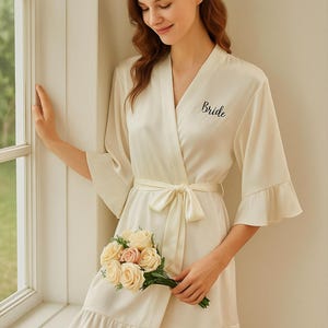 Könnte beinhalten: Elfenbeinfarbener Satin-Kimono mit Wickeldesign, Bindegürtel und Rüschensaum. Das Wort "Bride" ist auf der Vorderseite gestickt. Die Person hält einen Rosenstrauß. Ein Brautkimono.