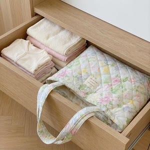 May include: A wooden drawer filled with folded baby clothes, towels, and a floral quilted tote bag. The tote bag features a diamond pattern and a pastel floral print with a small label. The drawer is open, revealing the contents.