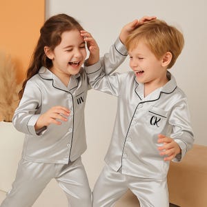 May include: Two children wearing matching silver pajamas with black trim and personalized initials. The pajamas have a button-down shirt and pants. The children are smiling and laughing.