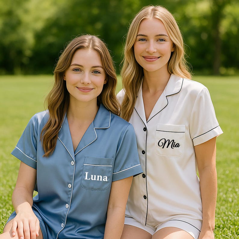 Op de afbeelding: Twee vrouwen dragen bijpassende pyjamasets. De ene set is lichtblauw met de naam "Luna" geborduurd op de zak. De andere set is wit met zwarte randen en de naam "Mia" geborduurd op de zak. Beide sets hebben korte mouwen en shorts.
