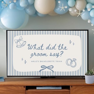 Könnte beinhalten: Ein TV-Schrank aus Holz mit einem Fernseher, auf dem der Text "What did the groom say?" angezeigt wird, ist mit einem blau-weißen Ballonbogen dekoriert. Eine kleine Topfpflanze und eine Kerze stehen auf dem Schrank.