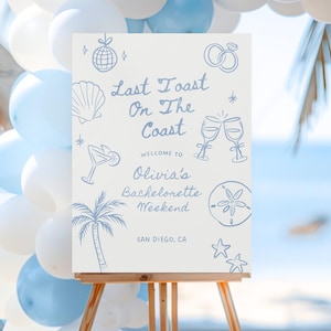 Puede incluir: Un letrero blanco sobre un caballete de madera con ilustraciones azules y el texto "Last Toast On The Coast" y "Olivia's Bachelorette Weekend, San Diego, CA". El letrero está decorado con globos.