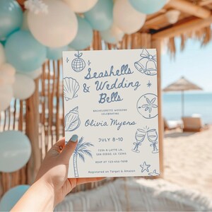 Peut inclure: Une invitation rectangulaire avec le texte "Seashells & Wedding Bells" en bleu, avec des illustrations de coquillages et de cloches. L'invitation est tenue devant une sc&egrave;ne de plage avec des ballons.