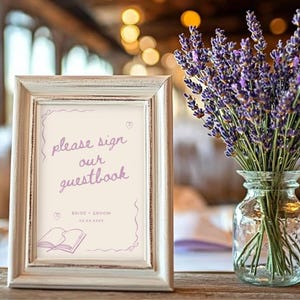 May include: A framed sign with the text "please sign our guestbook" in a soft purple script. The frame is distressed white, and a vase of lavender sits beside it. The scene is set on a wooden table, suggesting a wedding or event.