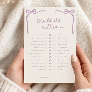 May include: A cream-colored game card with the text "Would she rather..." in a purple script font. The card features a list of questions with "or" options, and is decorated with purple bow accents. The card is held by a person.