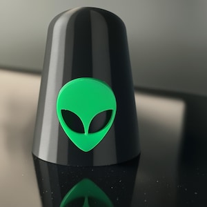 May include: A black, conical object with a bright green alien head emblem. The alien head is a classic design with large, almond-shaped eyes. The object has a smooth, glossy finish and is set against a dark background.