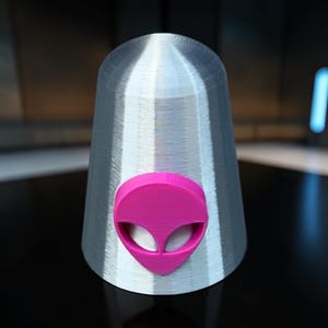 May include: A silver, cone-shaped object with a smooth, layered texture. Centered on the front is a raised, bright pink alien head emblem featuring two large, hollowed-out oval eye sockets and a pointed chin, creating a distinct contrast.