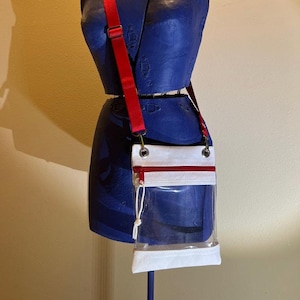 May include: A white crossbody bag with a clear front pocket and a red adjustable strap. The bag has a zipper closure and two silver eyelets.