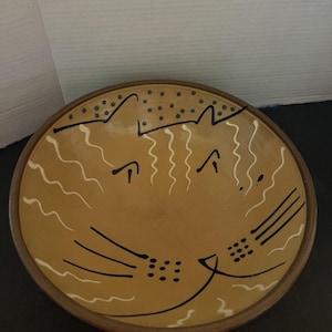May include: A round ceramic bowl with a tan glaze featuring a stylized cat face design. The interior is decorated with black painted whiskers, dots, and lines, along with white wavy lines. The bowl has a dark brown rim and a smooth, circular shape.