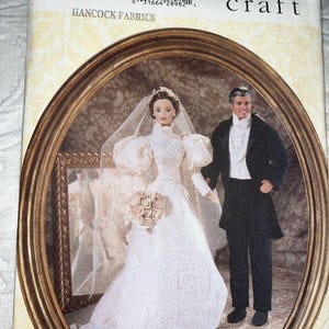 May include: A Vogue Craft 9985 sewing pattern envelope featuring an illustration of two dolls dressed in formal wedding attire. The bride wears a white gown with puffed sleeves and a veil, holding a bouquet, while the groom wears a black tuxedo.