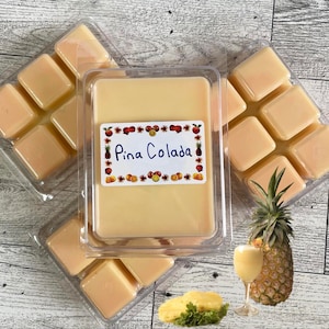 May include: Wax melts in clear plastic containers, with a label that reads "Pina Colada." The melts are a light yellow color, and the label has a decorative border of fruits and flowers. A pineapple and a glass of the drink are also in the image.